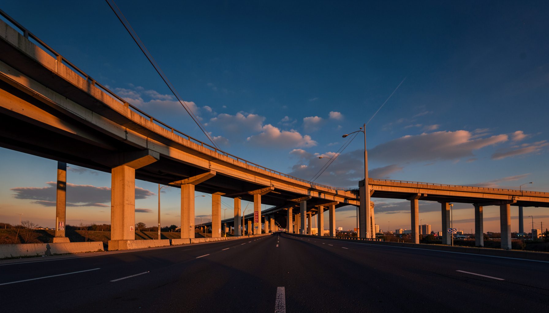 Wide-angle photograph of a sweeping highway overpass at golden hour, warm amber light casting long shadows across concrete pillars and asphalt, deep blue sky with soft clouds